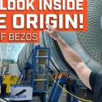 Hombre calvo con gafas de seguridad señalando un gran módulo esférico en el interior de una nave industrial dedicada a la fabricación de cohetes