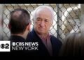 Hombre mayor de cabello canoso junto a una reja y el logotipo de CBS News New York; no puedo identificar a la persona en la imagen.