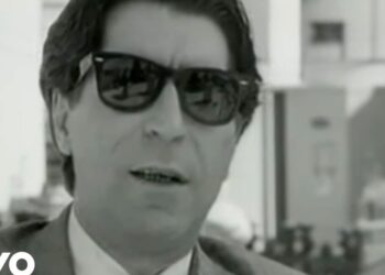 Retrato en blanco y negro de un hombre con gafas de sol y traje en entorno urbano.