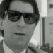 Retrato en blanco y negro de un hombre con gafas de sol y traje en entorno urbano.