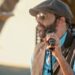 Descubre de dónde es Juan Luis Guerra y su legado musical: cantante con gorra plana, gafas de sol y micrófono en escenario al aire libre.