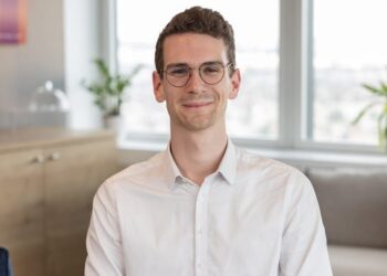 Joven profesional sonriente en oficina con camisa blanca y gafas, imagen ideal para contenido sobre Sueldo junior en KPMG: descubre cuánto puedes ganar y más.