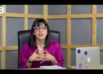 Mujer joven con gafas hablando sentada frente a un escritorio con portátil