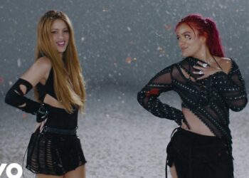 Dos artistas femeninas en escenario con atuendos negros y confeti, posando durante un video musical.