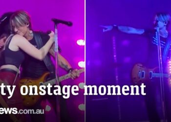 Cantante en el escenario abrazando a una fan con guitarra y luces púrpuras en un momento íntimo del concierto