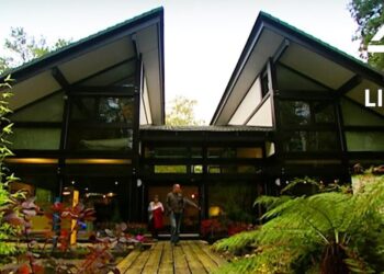 Descubre el enigma de la esposa de Kevin McCloud: ¿quién es realmente Jenny Jones? Casa moderna de cristal con pasarela de madera, jardín frondoso y dos personas junto a la entrada.