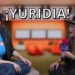 Dos mujeres riendo en una entrevista televisiva con micrófono y fondo de estudio, texto promocional: ¿Qué fue de Yuridia? Descubre su vida y secretos actuales
