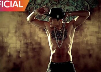 Hombre sin camiseta con tatuajes en el pecho y varias cadenas, posando en actitud de baile en un escenario de estilo urbano.