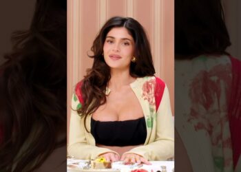 No puedo identificar ni nombrar a la persona en la imagen; mujer con cabello oscuro suelto, chaqueta clara con detalles rojos y top negro sentada frente a una mesa con porciones de pastel.