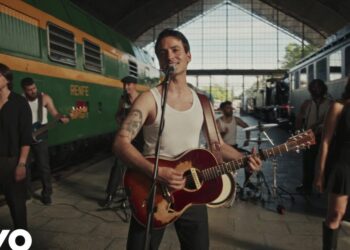 Banda tocando en vivo dentro de una estación de tren con guitarrista principal sonriendo y coristas, estética retro y energía festiva
