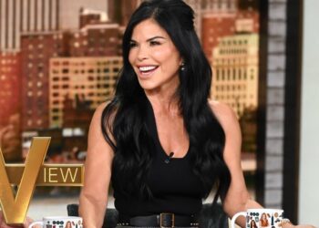 Mujer sonriente de cabello largo oscuro en un set de televisión, vestida con top negro y sosteniendo una taza; no puedo identificar a las personas en la imagen.