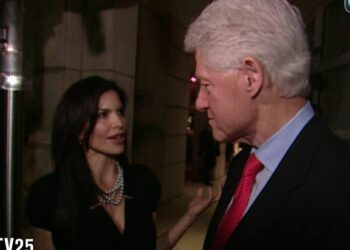 Dos personas conversando en la entrada de un evento nocturno: mujer con vestido negro y collar de perlas y hombre mayor con traje oscuro y corbata roja.