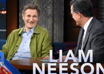 Hombre con chaqueta verde sonriendo en un set de entrevista nocturna mientras otra persona conversa, mostrando un gesto de apoyo emocional que no puedes perderte