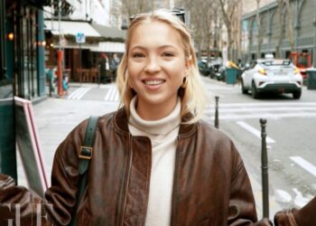 Mujer joven rubia sonriente con chaqueta de cuero marrón y jersey claro en una calle urbana