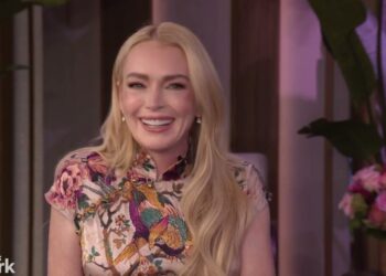 Mujer rubia sonriente en un set de entrevista con vestido floral y peinado suelto junto a un ramo de flores