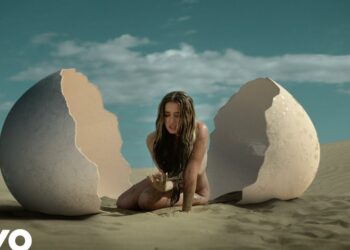 Mujer emergiendo de una cáscara de huevo gigante en un desierto bajo un cielo azul, lola indigo