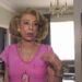 Mujer mayor de pie en un salón elegante con camiseta rosa y collar llamativo