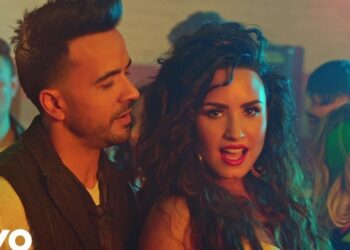 Primer plano de dos cantantes en un videoclip con iluminación cálida y ambiente de fiesta, no puedo identificar a las personas en la imagen.