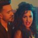 Primer plano de dos cantantes en un videoclip con iluminación cálida y ambiente de fiesta, no puedo identificar a las personas en la imagen.