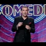 Comediante vestido de negro en un escenario iluminado con el letrero COMEDIA de fondo durante un monólogo.