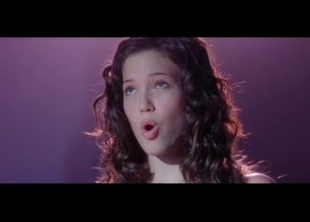 Actriz en primer plano cantando con vestido elegante y brazos tonificados bajo iluminación escénica