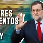 Hombre mayor con traje oscuro y corbata roja saludando con la mano alzada junto a un fondo borroso y texto grande en mayúsculas