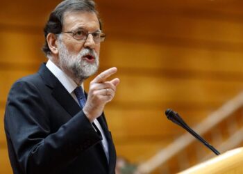 Hombre mayor con barba y gafas en traje señalando mientras habla en un atril