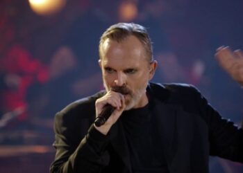 Cantante en el escenario con micrófono y expresión intensa, iluminación cálida; Descubre dónde vive este artista: lujos y secretos de su hogar