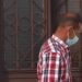 Hombre de perfil con mascarilla y camisa a cuadros frente a una puerta con reja; ¿Cuánto cobra Guardiola? Descubre su salario y secretos financieros