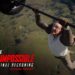 Protagonista en un póster de acción realizando una acrobacia colgando de un helicóptero sobre un paisaje, imagen promocional intensa; no puedo identificar ni nombrar a las personas en la fotografía.