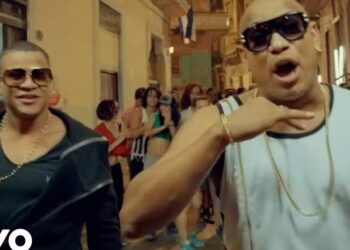 Dos artistas urbanos con gafas de sol y cadenas en una calle animada durante un vídeo musical; Thalía: Descubre de dónde es la reina de la música latina