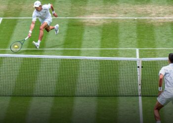 Dos tenistas en la red durante un punto sobre césped en Wimbledon, uno lanzándose para golpear la pelota mientras el rival espera al otro lado de la red.