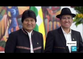 Dos hombres sonrientes con trajes tradicionales frente a una bandera multicolor en un retrato institucional.