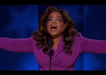 Mujer oradora en escenario con chaqueta morada, gafas y brazos extendidos delante de un micrófono