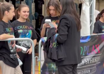 Aficionados con camisetas y merchandising junto a un puesto de venta en un concierto; Coldplay: descubre de dónde es la icónica banda británica.