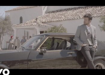 El sorprendente caché de Pablo López: ¿cuánto cobra por concierto? Hombre elegante apoyado en un coche clásico frente a una iglesia rural.