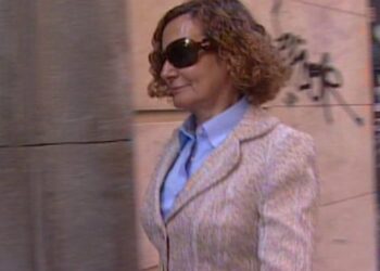 Mujer con abrigo claro y gafas de sol caminando por una calle urbana con graffiti en la pared