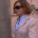 Mujer con abrigo claro y gafas de sol caminando por una calle urbana con graffiti en la pared