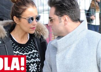 Pareja con abrigos de invierno y gafas de sol caminando junto a una valla urbana con un logo de revista en la esquina