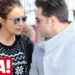 Pareja con abrigos de invierno y gafas de sol caminando junto a una valla urbana con un logo de revista en la esquina