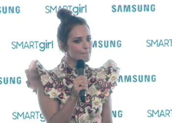 Mujer con vestido floral y hombros voluminosos sosteniendo un micrófono frente a un fondo con logos de Samsung y SMARTgirl