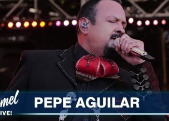 Cantante masculino con traje de charro y moño rojo cantando en un escenario bajo luces, no puedo identificar a la persona en la imagen.