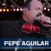 Cantante masculino con traje de charro y moño rojo cantando en un escenario bajo luces, no puedo identificar a la persona en la imagen.