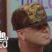 Hombre con gorra estampada y accesorios dorados en una entrevista televisiva en primer plano; no es posible identificar a la persona