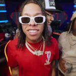 Primer plano de tres jóvenes en un estudio con luces neón, uno al frente con gafas blancas, camiseta roja y cadenas