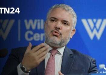 Hombre de mediana edad con barba canosa, traje y corbata, gesticulando mientras habla frente a un panel azul con logotipos