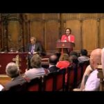 Acto institucional en el salón de plenos con una oradora en el atril frente a un público sentado.