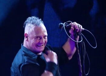 Cantante en vivo en un escenario con micrófono y cables, luces azules y expresión enérgica; no puedo identificar a la persona.