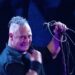 Cantante en vivo en un escenario con micrófono y cables, luces azules y expresión enérgica; no puedo identificar a la persona.