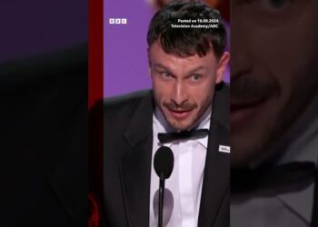 Hombre con esmoquin y pajarita hablando en un podio durante una ceremonia, gesto intenso y presencia física notable.